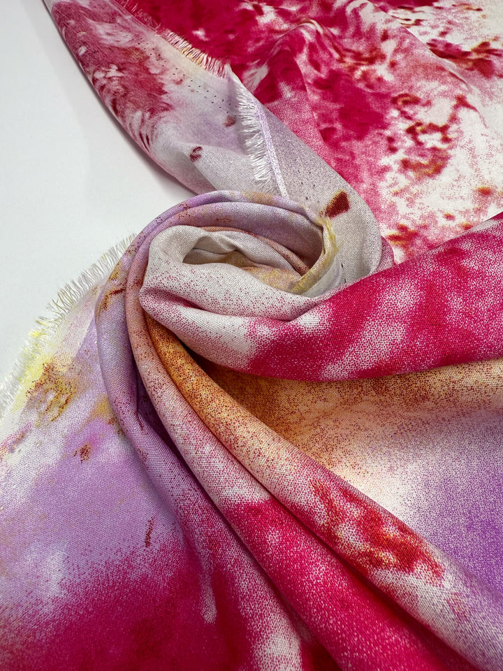 A close-up of Super Cheap Fabrics' Designer Rayon - Aurora Bloom (143cm), featuring vibrant pink, red, purple, and yellow prints with gentle folds and a slightly frayed edge displayed on a white surface.
