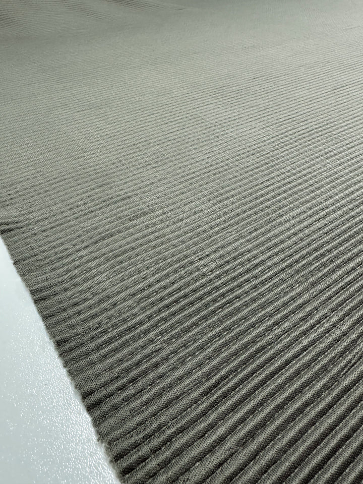 The close-up shows the ribbed, textured surface of Pleat - Pewter - 140cm by Super Cheap Fabrics. The gray polyester fabric fills the frame with evenly spaced parallel ridges and a contrasting matte finish on the smoother left edge.