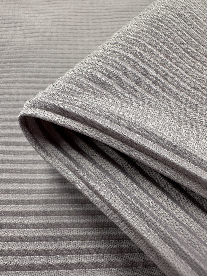 Close-up of folded gray ribbed polyester fabric shows horizontal ridges with an alternating pattern of raised and flat areas. Despite its soft, thick look, this lightweight fabric, Pleat - Violet Ice - 140cm by Super Cheap Fabrics, is suitable for warm clothing or upholstery.
