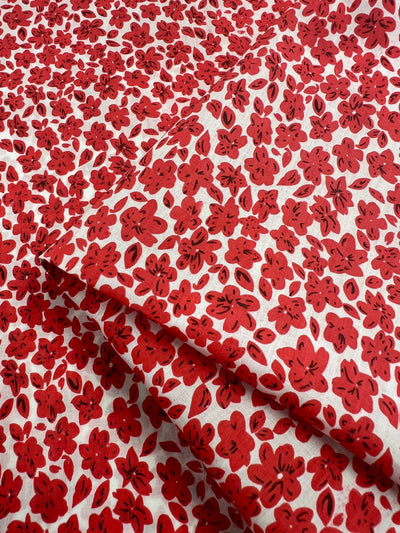 A folded 100% cotton fabric known as "Printed Cotton - Red Rain - 145cm" from Super Cheap Fabrics, featuring a vibrant pattern of red flowers with black accents on a white background. The dense floral design covers the entire surface of this lightweight, printed cotton fabric.