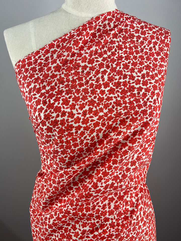 The Super Cheap Fabrics' "Printed Cotton - Red Rain - 145cm" draped over a mannequin features a bold red and white floral pattern. The lightweight 100% cotton fabric covers one shoulder and crosses over the body, creating a visually striking design. The neutral background highlights the vibrant colors of this fabric.