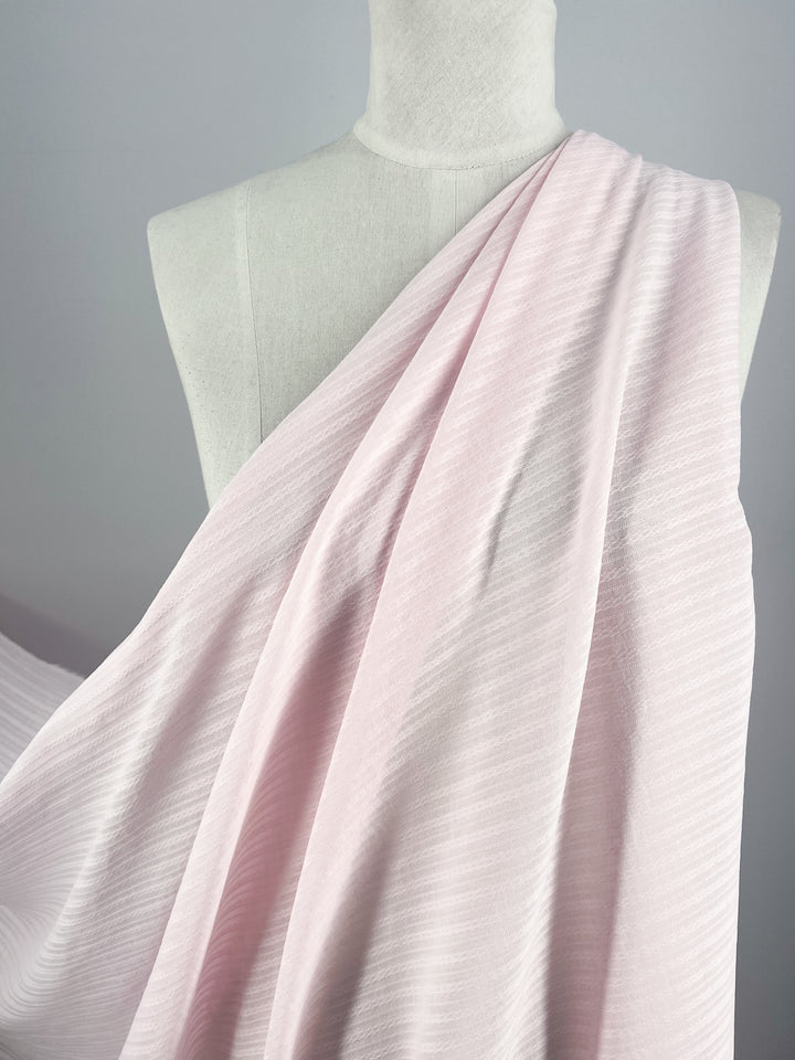 A mannequin draped with Textured Cotton - Shrinking Violet from Super Cheap Fabrics, showcasing its soft pink hue in an extra light weight 100% cotton fabric with subtle diagonal stripes. The 140cm wide material flows gracefully over the shoulders, highlighting its delicate texture and smooth drape against a neutral background.