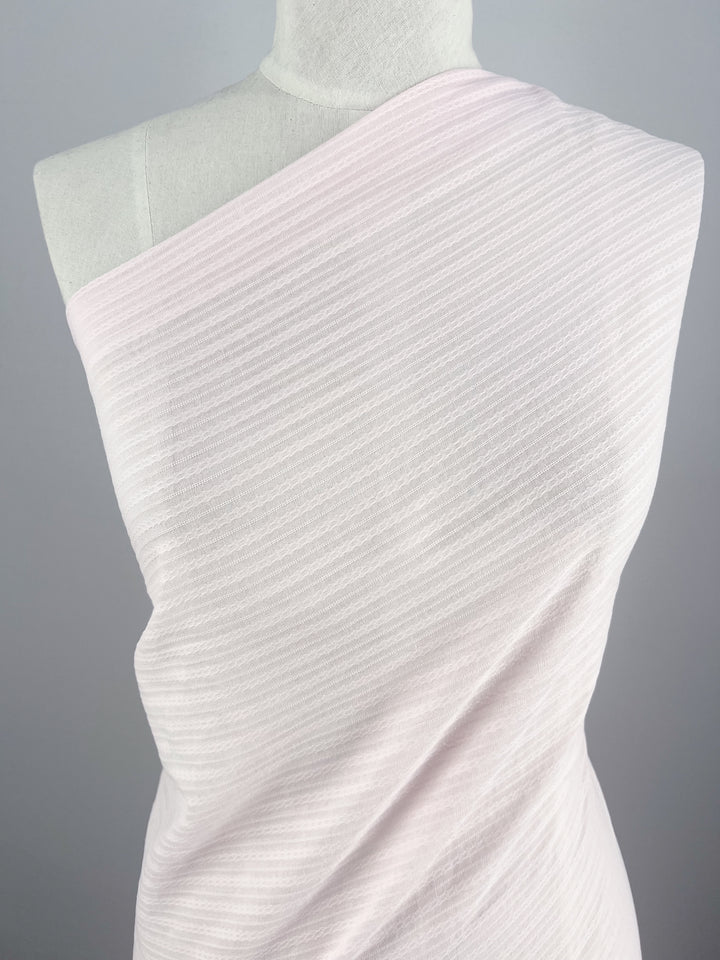 A mannequin draped with Textured Cotton - Shrinking Violet - 140cm by Super Cheap Fabrics, an extra light-weight fabric in soft, light pink featuring subtle horizontal stripes. The 100% cotton fabric is wrapped asymmetrically over one shoulder, revealing its texture and elegant drape. The background is a plain light gray.