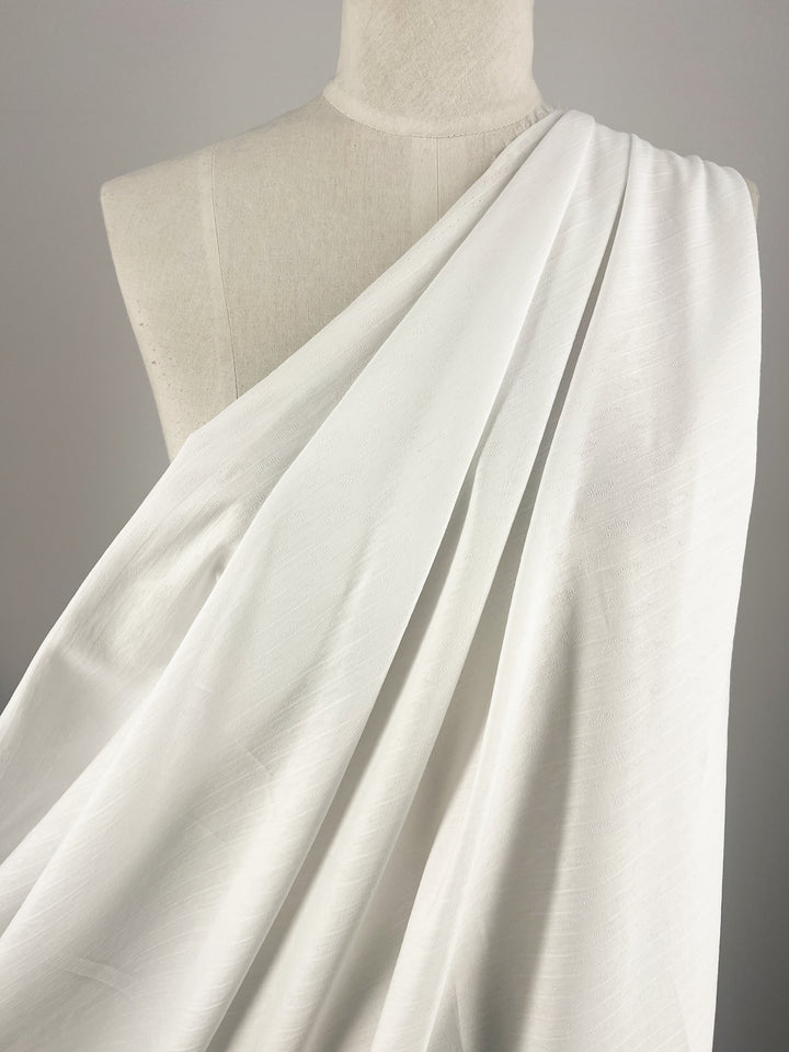 A close-up of a mannequin draped in Super Cheap Fabrics' Textured Cotton - White - 145cm. The material appears soft and smooth, flowing naturally over the mannequin's shoulder in a classic, elegant manner. The background is plain, keeping the focus on the beautifully textured fabric.