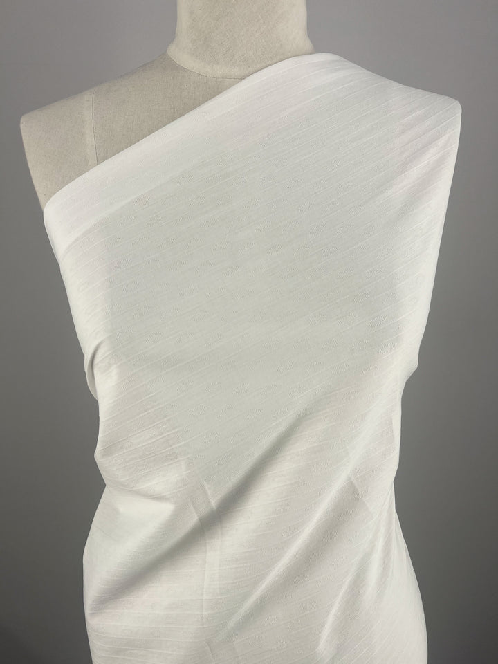 A torso mannequin wrapped in a single-shoulder white fabric from Super Cheap Fabrics' Textured Cotton - White - 145cm collection drapes gracefully across the chest and torso. The 100% cotton fabric appears lightweight and slightly textured, with subtle vertical lines. The plain gray background accentuates the simplicity and elegance of the draped fabric textures.