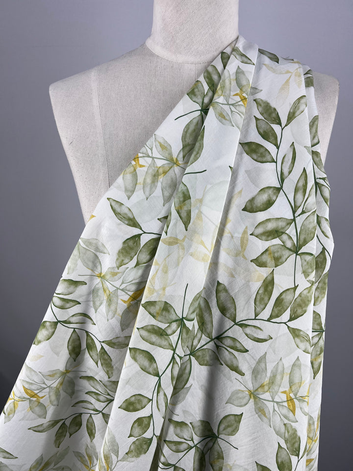 The Designer Cotton - Gully - 150cm from Super Cheap Fabrics, featuring a lightweight cotton fabric with a green leafy pattern, is draped over a dress form. The design showcases shades of green leaves in varying hues, creating a natural, botanical appearance. Perfect for luxury sewing projects, the display is set against a neutral gray background.