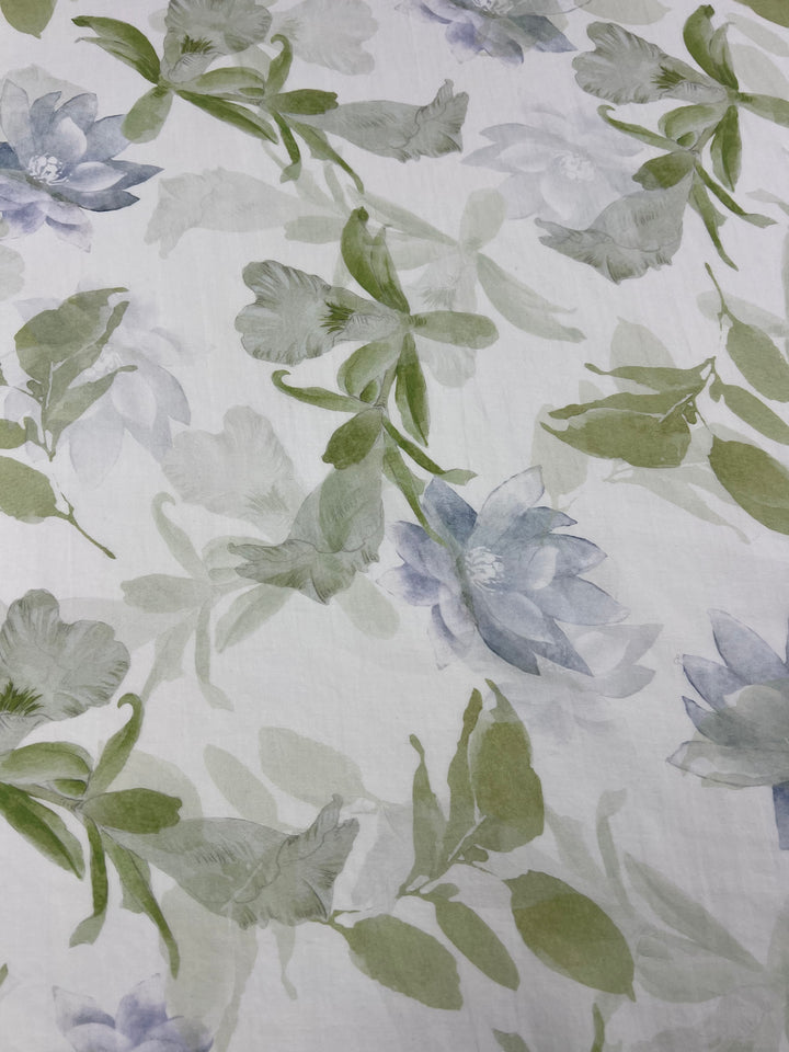 A close-up of the Super Cheap Fabrics 'Designer Cotton - Lotus - 150cm' showcases a stunning watercolor floral pattern with blue and green flowers and leaves on a white background. Perfect for luxury sewing projects, this fabric features soft, delicate appearances with varying shades of green foliage interspersed among the blossoms.