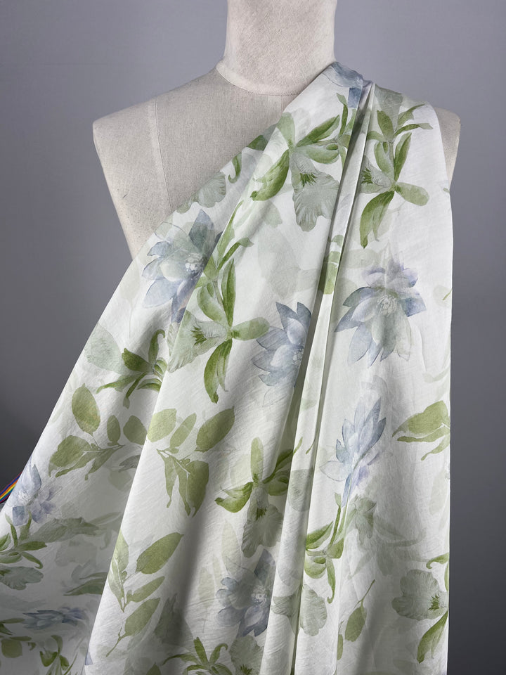 A white dress form draped with Super Cheap Fabrics' Designer Cotton - Lotus - 150cm, showcasing a delicate pattern of green leaves and light blue flowers. The minimalist backdrop highlights the soft, elegant material—perfect for luxury sewing projects.