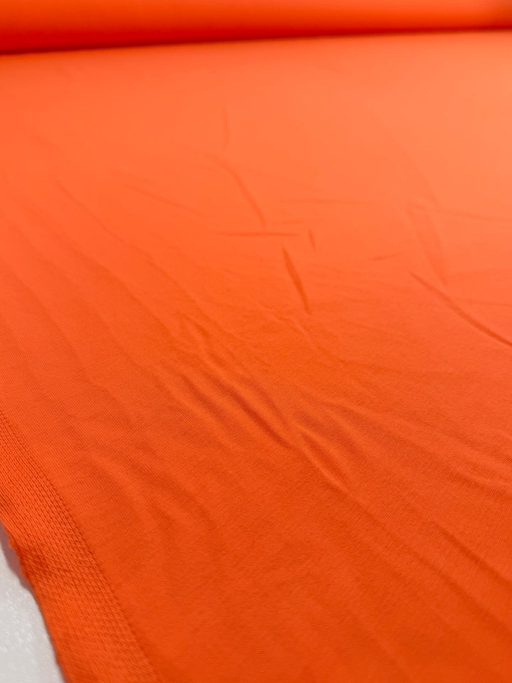 A close-up of Super Cheap Fabrics’ Cotton Jersey - Mandarin Orange (165cm), showing visible creases and slight wrinkles as the bright fabric is unrolled on a flat surface.