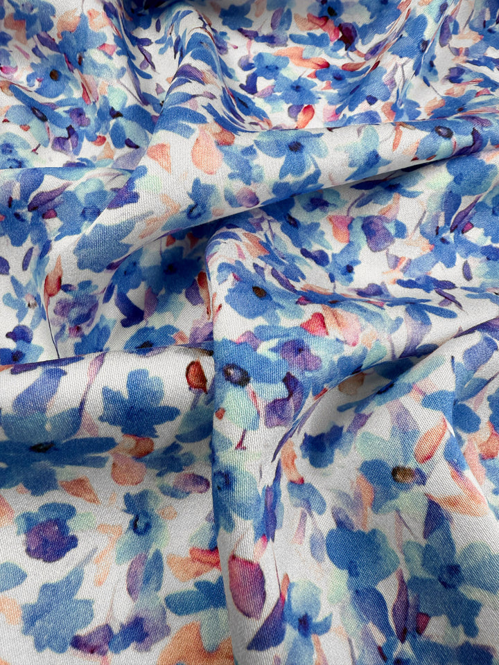 A close-up of Super Cheap Fabrics’ Mercerised Rayon - Bluebell Drift (150cm), showing soft, breathable material with blue, purple, and peach watercolor-style floral print on white, gently draped to reveal folds and shadows.