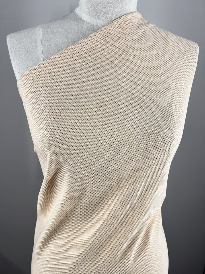 A dress form draped with Super Cheap Fabrics' Waffle Knit - Sun Kiss (170cm) features a beige, one-shoulder textured fabric with a soft honeycomb pattern, creating a three-dimensional look against the solid gray background.