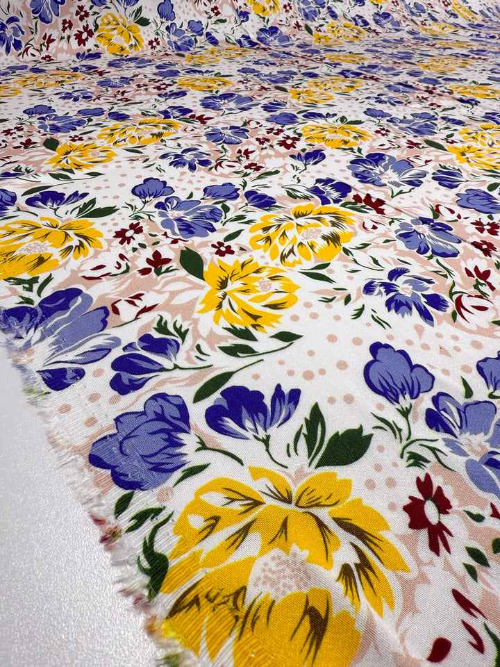 A close-up of Super Cheap Fabrics' Mercerised Rayon - Spring Bouquet (150cm), showing its breathable, lightweight material with vibrant yellow, blue, and red flowers with green leaves on white; the edge is slightly frayed in the foreground.