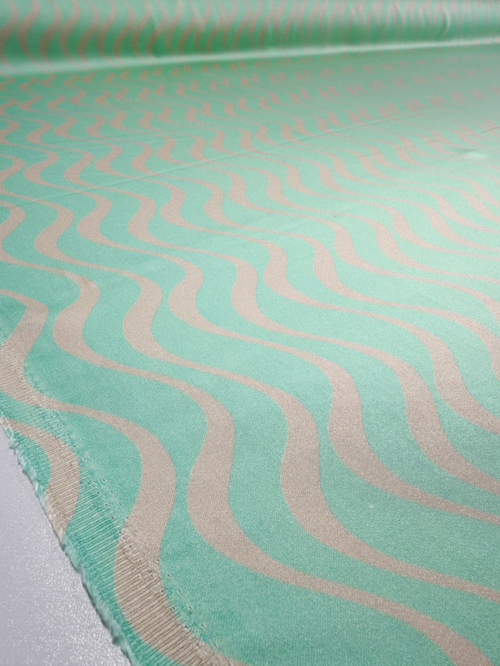 A roll of Super Cheap Fabrics’ Mercerised Rayon - Seafoam Rhythm (150cm) in mint green with a repeating wavy beige pattern is spread out flat, displaying its lightweight texture and unfinished edge.