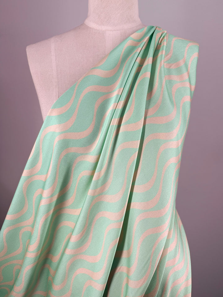 A dress form draped with Super Cheap Fabrics' Mercerised Rayon - Seafoam Rhythm (150cm), a breathable, lightweight fabric in mint green with a wavy cream pattern, arranged over one shoulder to suggest a toga or dress design.