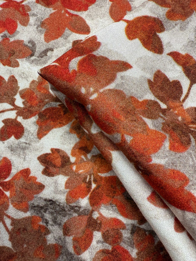A close-up of Super Cheap Fabrics' Mercerised Rayon - Crimson Canopy (150cm), showing its lightweight, breathable material with a red and orange autumn leaf pattern on a white background, slightly folded.