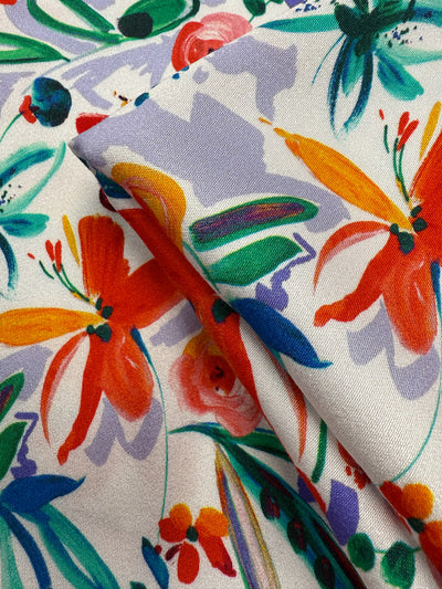 Close-up of Super Cheap Fabrics' Mercerised Rayon - Tropical Brushstroke (150cm) showing bright orange, yellow, green, and pink floral patterns with leaves on a white background. The lightweight fabric is partially folded to reveal its vibrant texture.