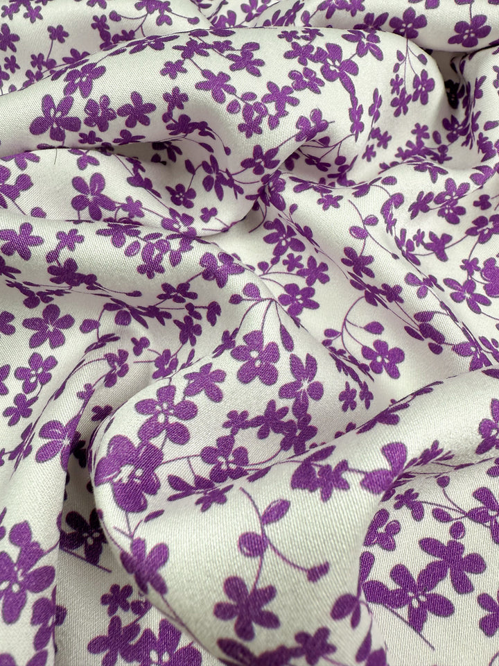 Super Cheap Fabrics' Mercerised Rayon - Violet Meadow - 150cm features white, breathable fabric with a scattered purple floral pattern. The lightweight, slightly wrinkled material forms gentle folds and soft shadows across its surface.