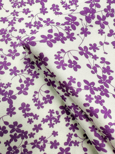 A close-up of Super Cheap Fabrics’ Mercerised Rayon - Violet Meadow (150cm) shows its lightweight, breathable fabric in a small violet floral and vine pattern, with a visible crease formed by a fold.
