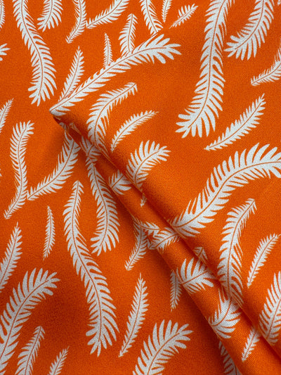 Super Cheap Fabrics’ Mercerised Rayon - Citrus Flow - 150cm is an orange fabric with a white leaf or feather pattern, showing overlapping sections that highlight the repeating design and its fluid drape.