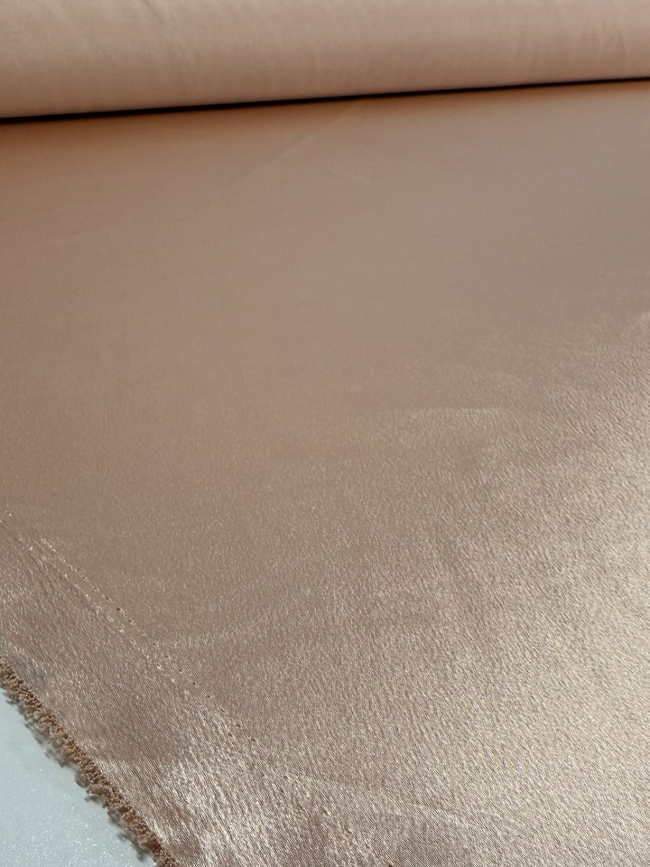 A close-up of Super Cheap Fabrics’ Satin Back Crepe in Cream Tan (150cm), showing its shiny, smooth texture as it’s partially unrolled on a flat surface.