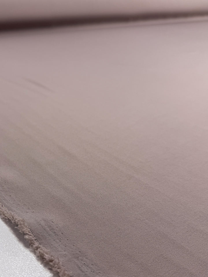 A close-up of Super Cheap Fabrics' Liquid Twill - Goat (150cm), a smooth, light brown suiting fabric with a slightly frayed edge. Its soft, even texture is highlighted by subtle shadows and gentle creases.