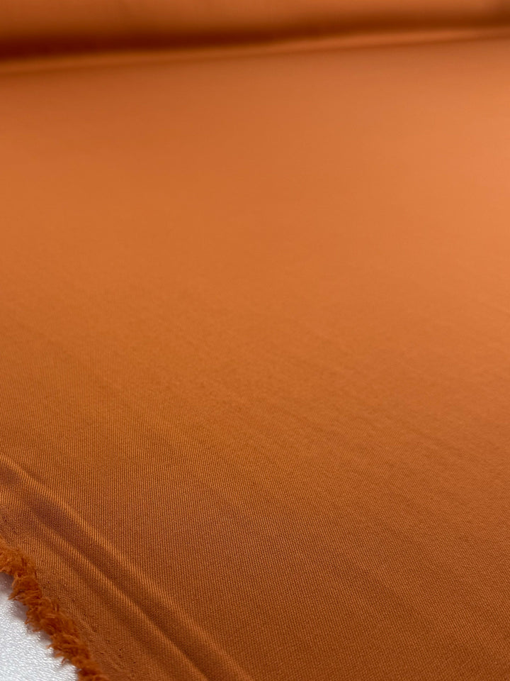 A close-up of Super Cheap Fabrics' Liquid Twill - Tomato Cream (150cm) shows the smooth, rust-orange polyester fabric laid flat with a slightly frayed left edge, highlighting its rich color and texture.