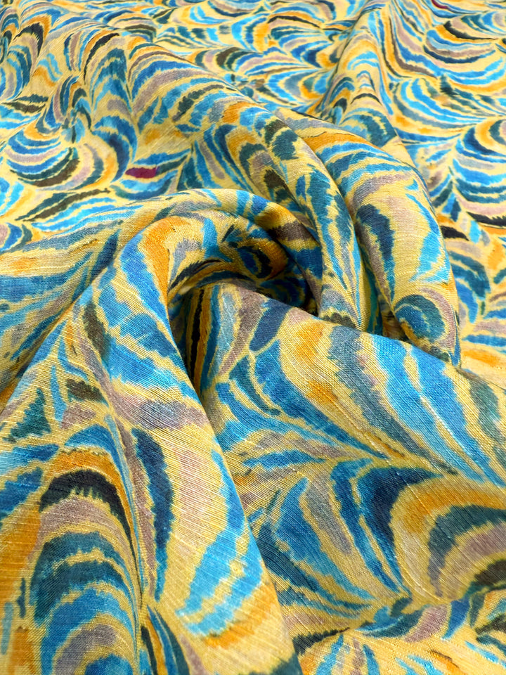 A close-up of Super Cheap Fabrics’ Textured Designer Cotton - Spiral Sun (145cm), showing its vibrant yellow, blue, and brown marbled swirls with elegant folds that highlight the fabric’s texture.