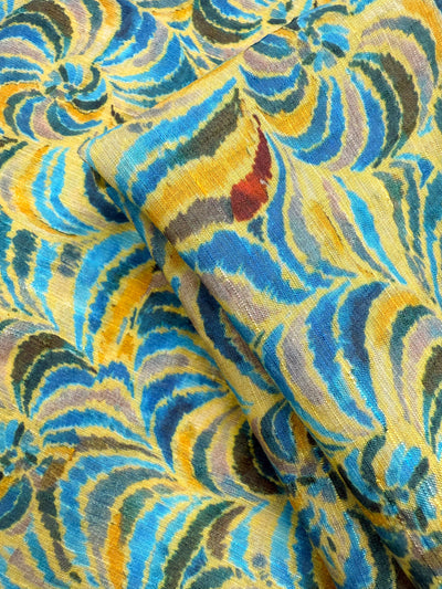A close-up of Super Cheap Fabrics' Textured Designer Cotton - Spiral Sun (145cm), featuring a lightweight, colorful fabric with swirling abstract patterns in yellow, blue, brown, black, and red; one piece folded over another.