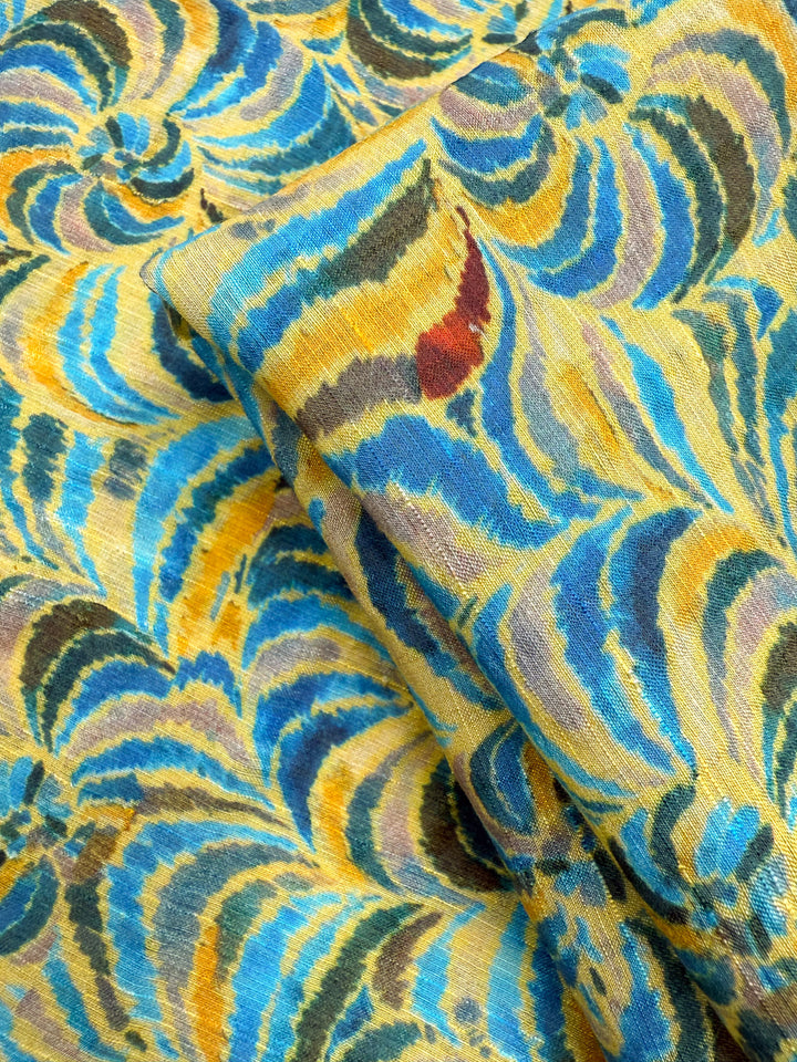 A close-up of Super Cheap Fabrics' Textured Designer Cotton - Spiral Sun (145cm), featuring a lightweight, colorful fabric with swirling abstract patterns in yellow, blue, brown, black, and red; one piece folded over another.