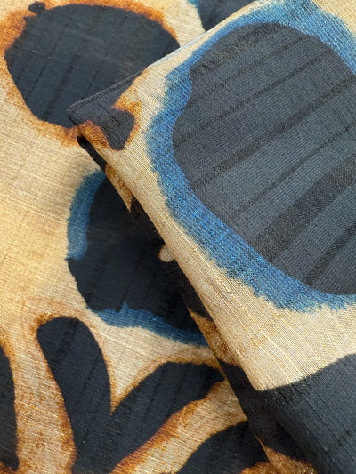 A close-up of Super Cheap Fabrics' Textured Designer Cotton - Terra Dot (145cm), featuring two overlapping pieces with a bold black, blue, and tan abstract pattern on a textured beige background. The fabric looks soft and slightly sheer.