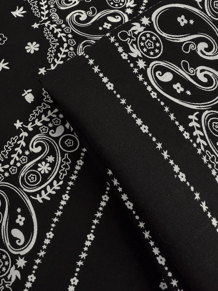 Close-up of Designer Rayon - Bandana Noir (148cm) by Super Cheap Fabrics: black lightweight rayon with a white paisley and floral bandana pattern, featuring intricate swirls, flowers, and dotted lines.