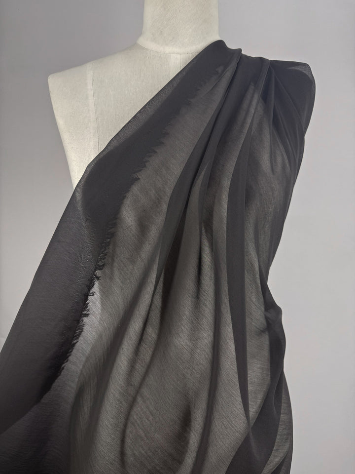 A mannequin is draped with Super Cheap Fabrics' Designer Voile - Black - 150cm, the sheer, lightweight fabric elegantly cascading over one shoulder and softly revealing the mannequin's contours against a plain gray background.