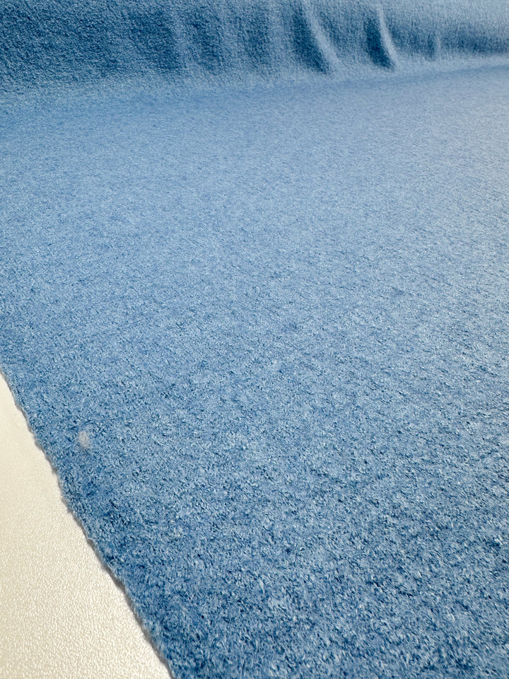 A close-up of "Boiled Wool - Allure - 150cm" from Super Cheap Fabrics shows a blue wool-viscose blend fabric spread flat with subtle folds creating a wave effect. The fabric appears soft and smooth, and its edge is visible on the left against a white surface.