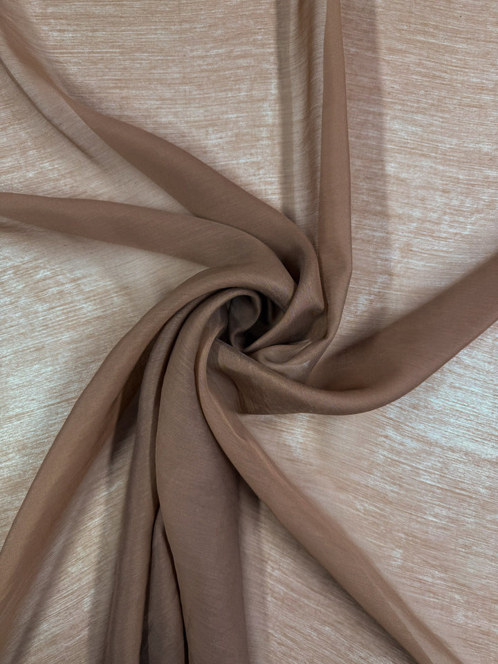 A close-up of Super Cheap Fabrics' Designer Voile - Burro - 150cm shows sheer, light brown fabric twisted into a spiral, with soft folds and a gentle play of light and shadow on its textured surface.