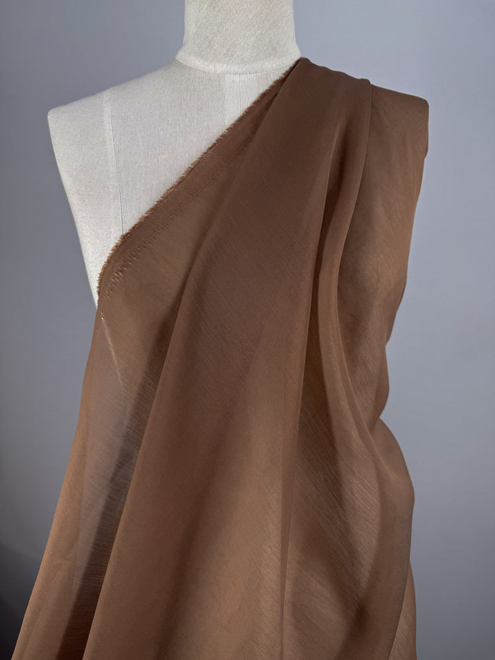 A mannequin showcases Super Cheap Fabrics’ Designer Voile - Burro - 150cm, an ultra-lightweight sheer fabric in light brown, draped over one shoulder and set against a plain gray background.