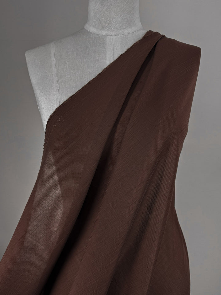 A mannequin is draped in Super Cheap Fabrics' Textured Designer Voile - French Roast (155cm), wrapped diagonally across one shoulder and set against a plain gray background.
