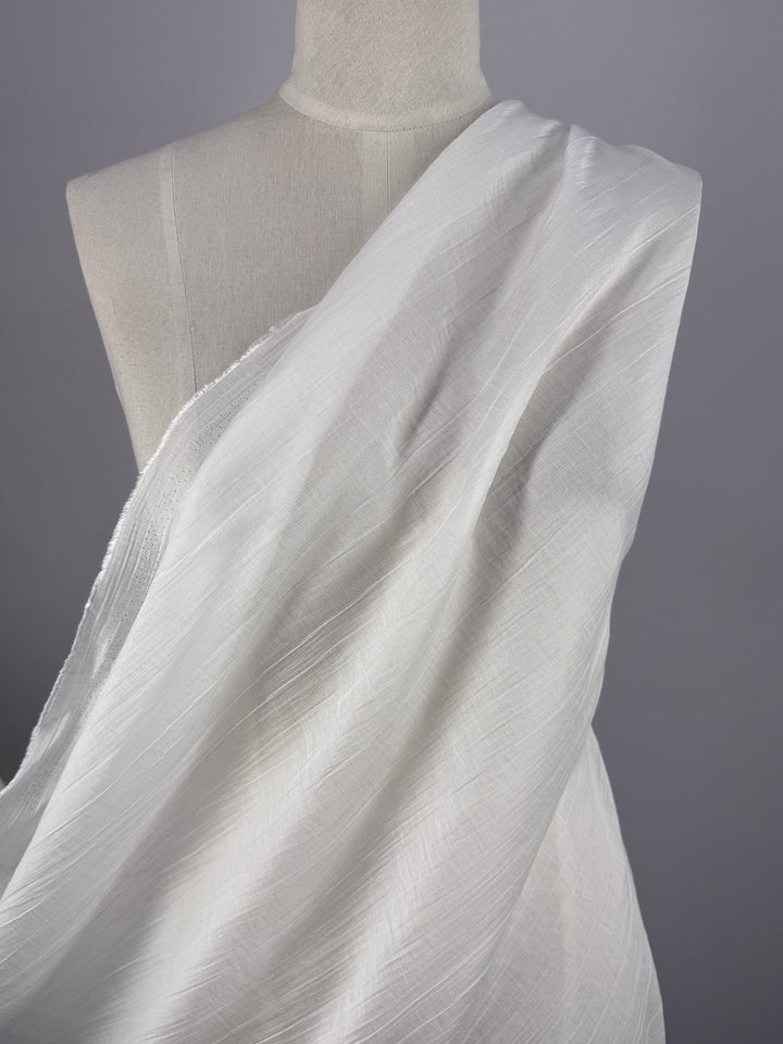 A close-up of Super Cheap Fabrics’ Textured Designer Voile - Ivory - 155cm draped over a dress form, covering one shoulder and creating soft, flowing folds against a neutral gray background.