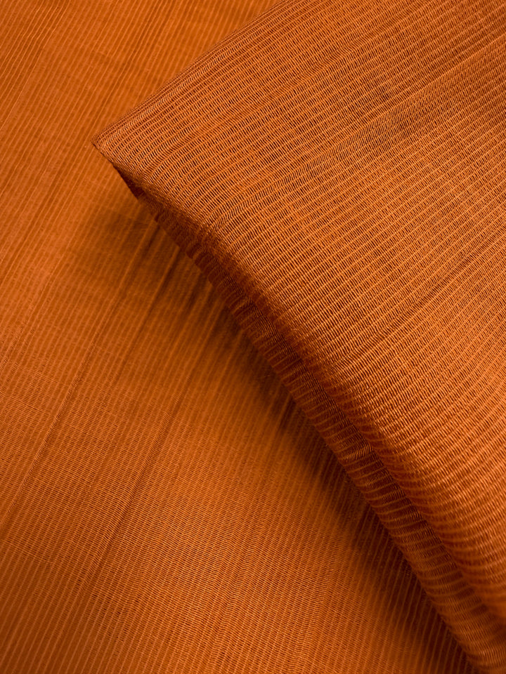 Close-up of Super Cheap Fabrics’ Textured Designer Voile in Rust (155cm): an orange cotton-nylon blend with fine parallel lines, smooth texture, and a detailed weave, shown with one layer neatly folded over another.