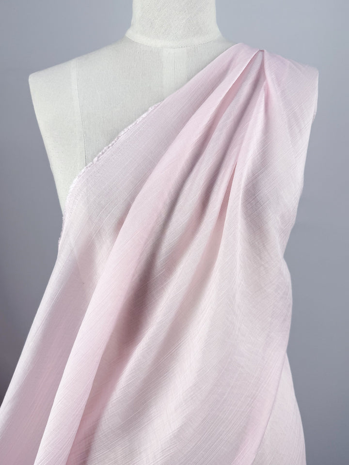 A mannequin displays Super Cheap Fabrics' Textured Designer Voile - Carinaria (155cm), a soft, light pink, semi-sheer fabric with a subtle texture, draped over one shoulder against a plain gray background.
