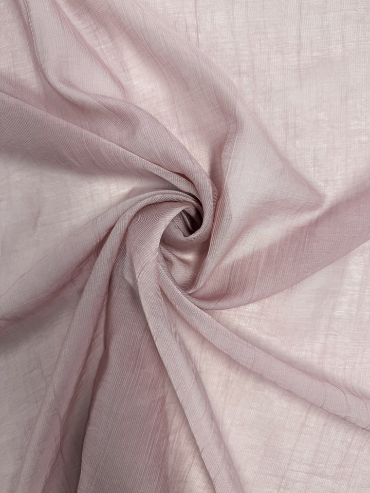 A close-up of Super Cheap Fabrics' Textured Designer Voile - Silver Pink (155cm), showcasing its sheer, lightweight, softly crinkled texture arranged in spiral folds—ideal for summer dresses.