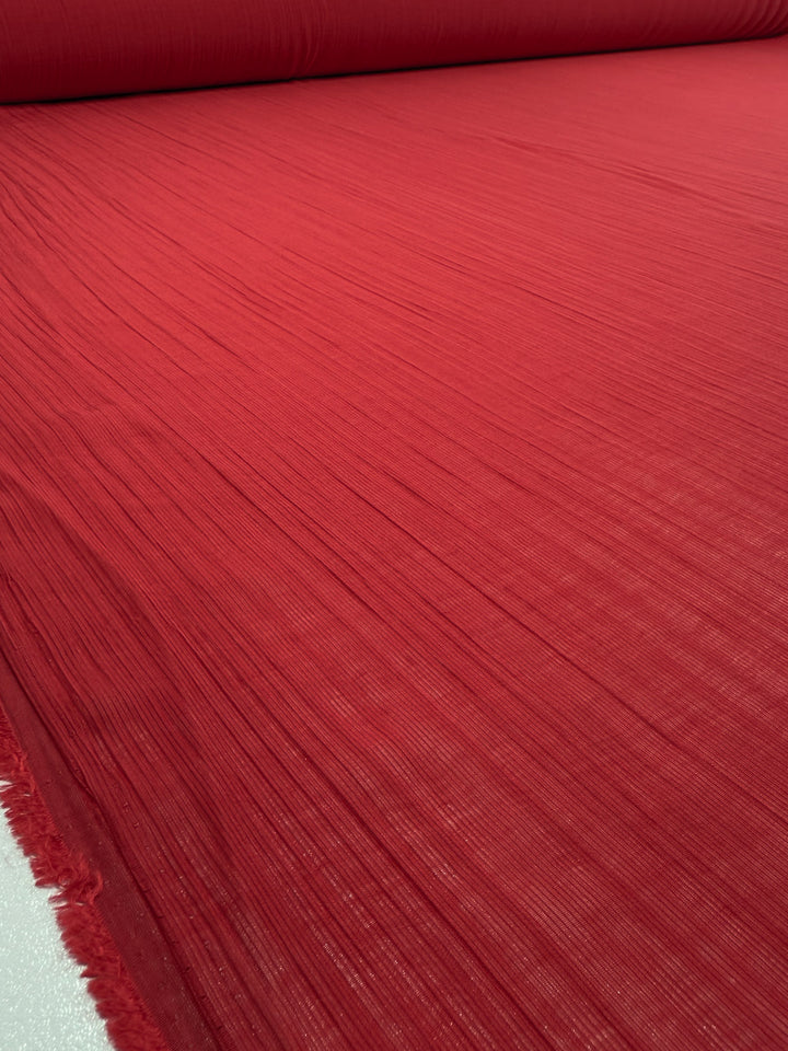 A close-up view of Super Cheap Fabrics' Textured Designer Voile - True Red (155cm), showing visible vertical lines and a semi-sheer, unrolled fabric across a flat surface.