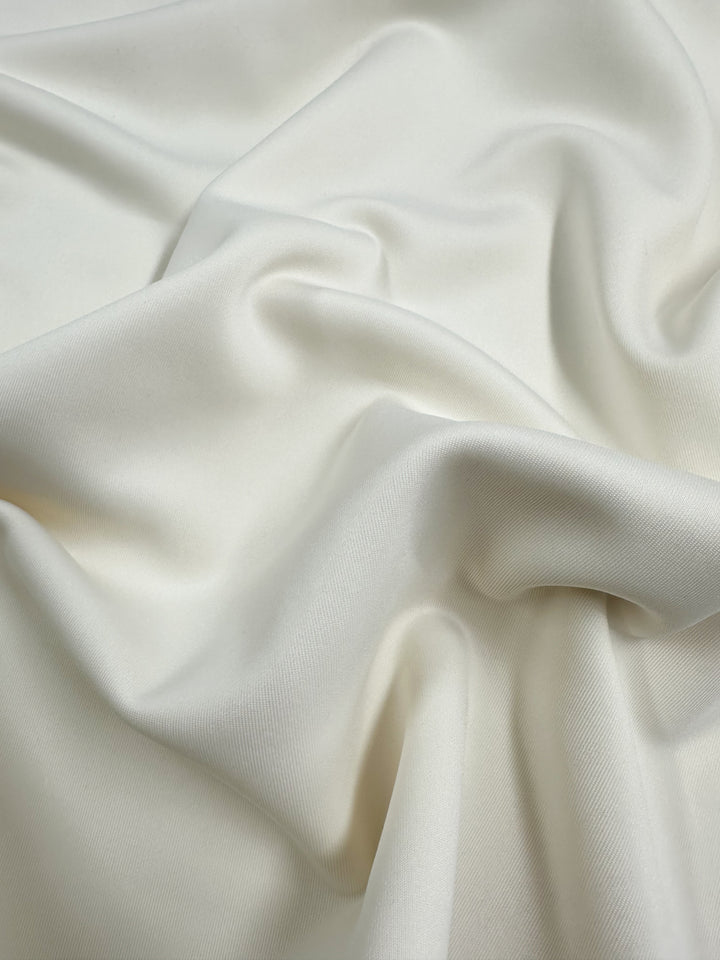 A close-up view of the Milano Ponte - Ivory fabric by Super Cheap Fabrics reveals its light beige, soft, and smooth texture with gentle folds and drapes that create flowing highlights, reminiscent of medium to heavy weight satin.