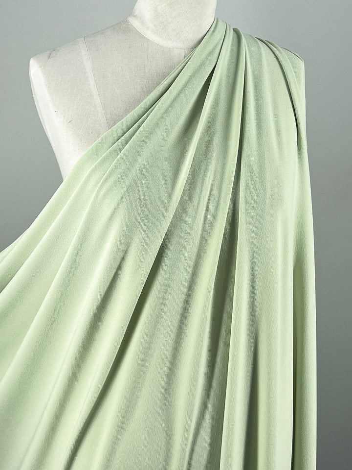 The pistachio Crepe Lycra fabric from Super Cheap Fabrics elegantly cascades over a mannequin on a gray background. At 148cm wide, this soft, medium-weight fabric flows smoothly across the form.