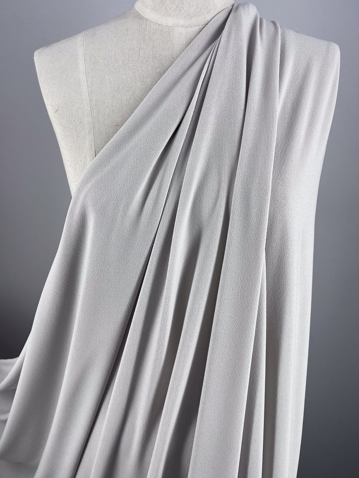 A close-up of a draped piece of Crepe Lycra - Silver - 148cm by Super Cheap Fabrics on a mannequin. The medium-weight fabric is arranged in folds, showcasing its texture and fluidity against the backdrop of a plain, neutral wall.