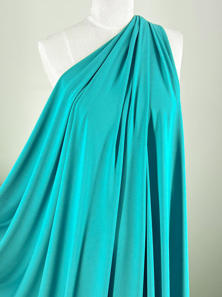 A mannequin displays a graceful, one-shoulder dress made from Super Cheap Fabrics' Crepe Lycra - Jade - 148cm, with an elegant drape and smooth medium weight texture featuring 94% polyester.