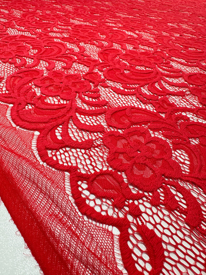 A close-up of Super Cheap Fabrics' Designer Lace - Scarlet - 145cm reveals an intricate floral pattern. This lace, ideal for evening dresses, showcases detailed flowers and leaves with a delicate design on a contrasting background.