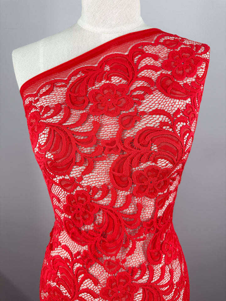 A mannequin displays Super Cheap Fabrics' Designer Lace - Scarlet - 145cm, showcasing its vibrant scarlet hue and intricate floral pattern. The sheer lace, perfect for evening dresses, is styled asymmetrically over one shoulder for an elegant look.