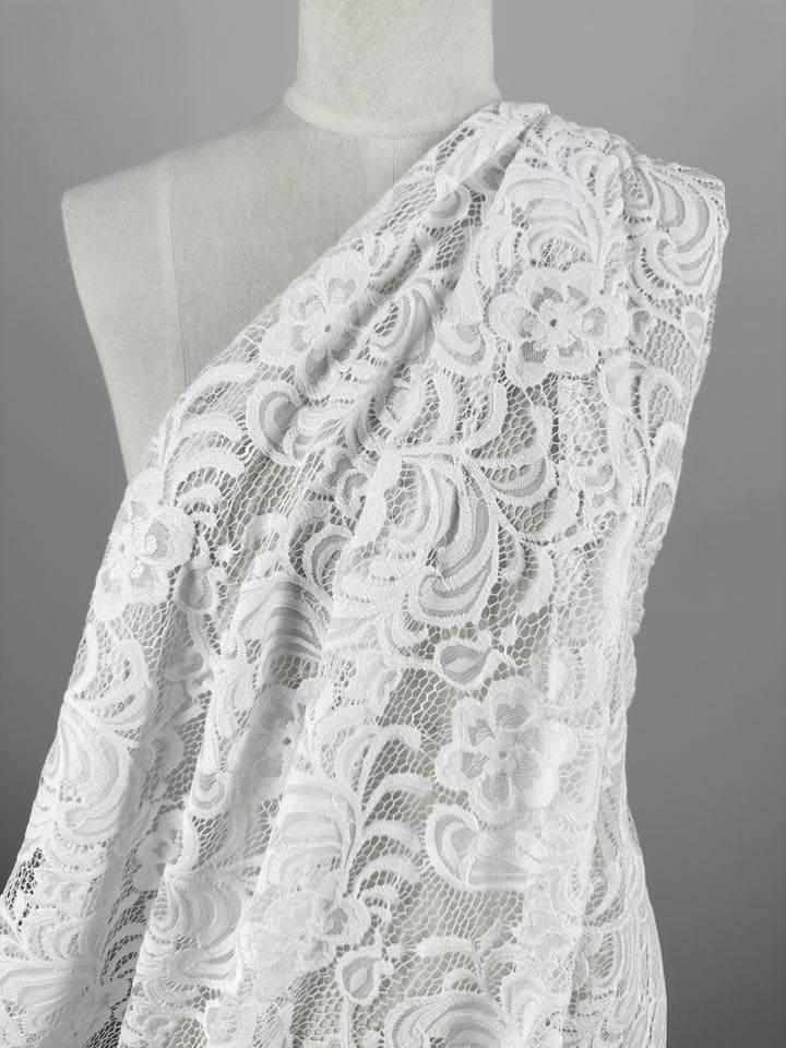 The Designer Lace - White - 145cm by Super Cheap Fabrics elegantly drapes on a mannequin, perfect for evening dresses. Its intricate floral and swirling patterns flow over one shoulder, showcasing the delicate texture against a plain gray background.