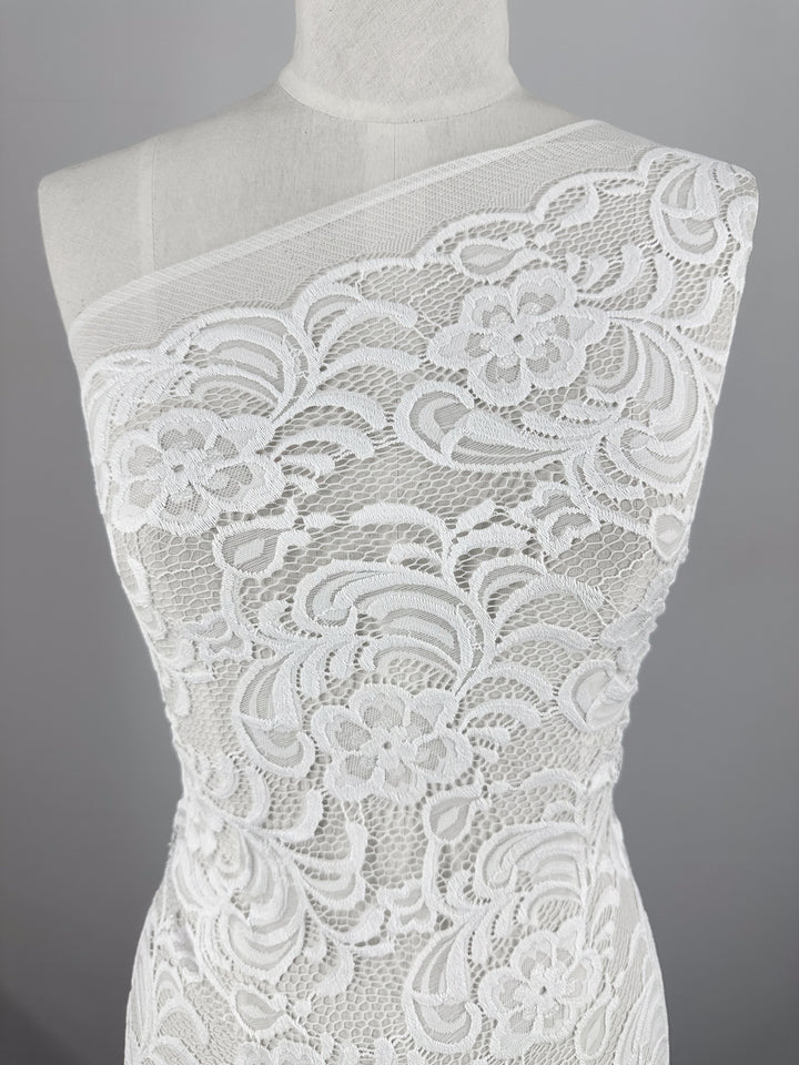 A close-up of Designer Lace - White - 145cm by Super Cheap Fabrics on a one-shoulder dress form showcases its floral and swirl patterns, ideal for elegant evening dresses. The neutral gray background accentuates the intricate texture and detail of the fabric.