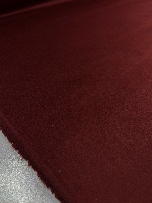 A close-up shot of the smooth, dark red Wool Cashmere - Fired Brick from Super Cheap Fabrics, showcasing its soft texture and slightly frayed edge on the left. The out-of-focus background draws attention to the rich color and luxurious texture of this 150cm fabric, making it an ideal choice for outer coats.